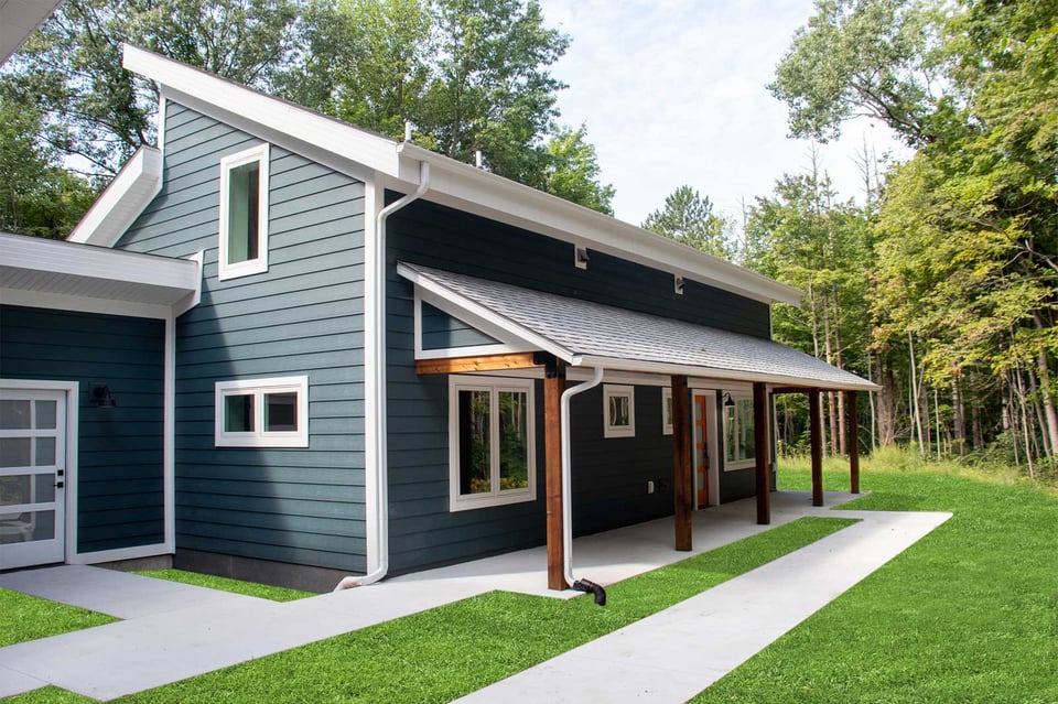 Modern blue two-story home exterior in West Michigan with white trim, wood posts, and forested backdrop