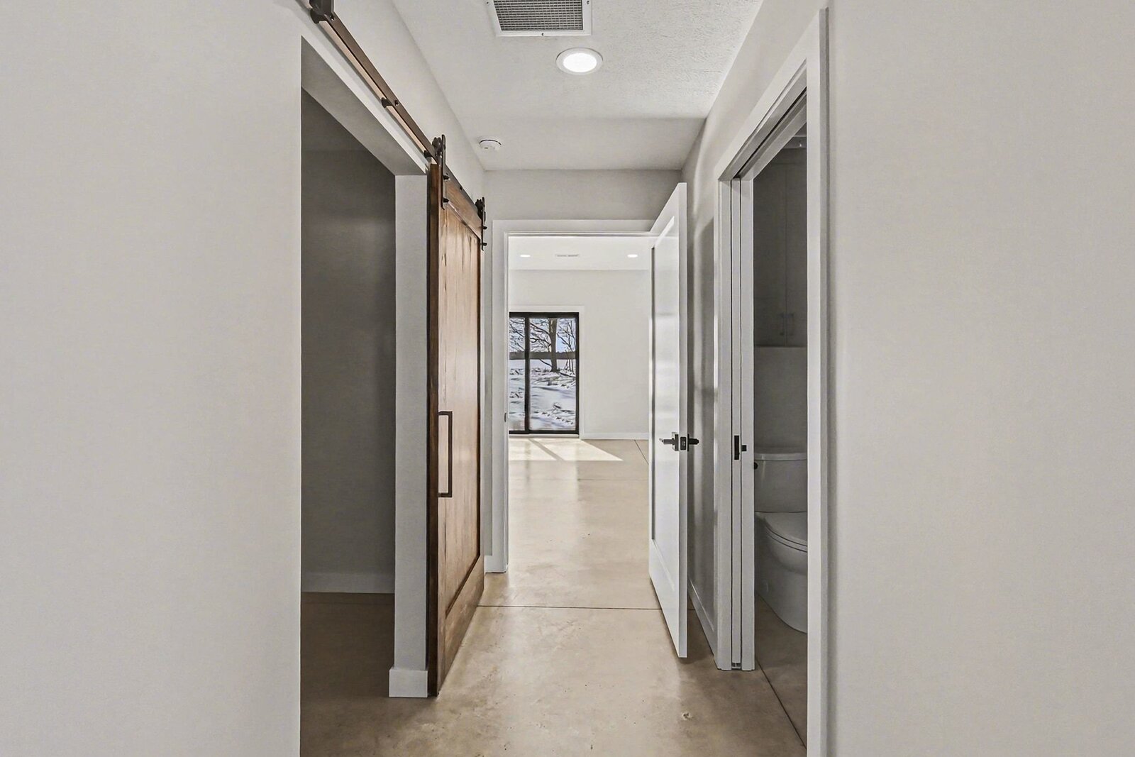Hallway in a modern Western Michigan home with polished concrete floors, sliding barn door, and view toward a bright room with snowy landscape outside
