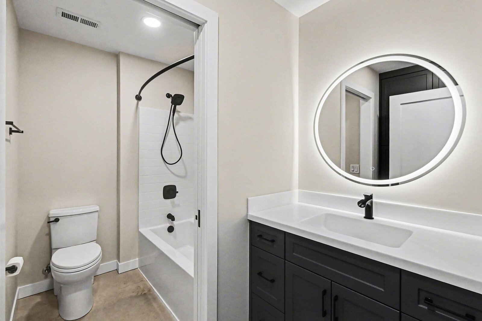 Modern bathroom in a Western Michigan home with tub shower combo, black fixtures, round lighted mirror, and dark vanity